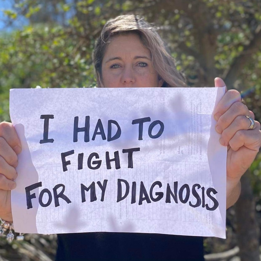 A woman with blonde hair holds up a sign to the camera about fighting for a diagnosis. 