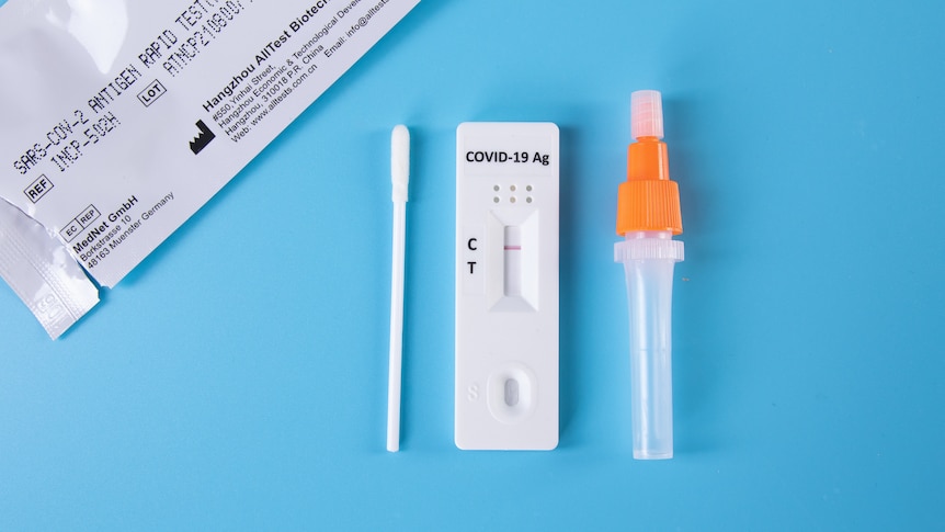 A generic image of a Rapid Antigen Test packet, a swab, a small dropper bottle, and the test itself laid out on a blue surface.