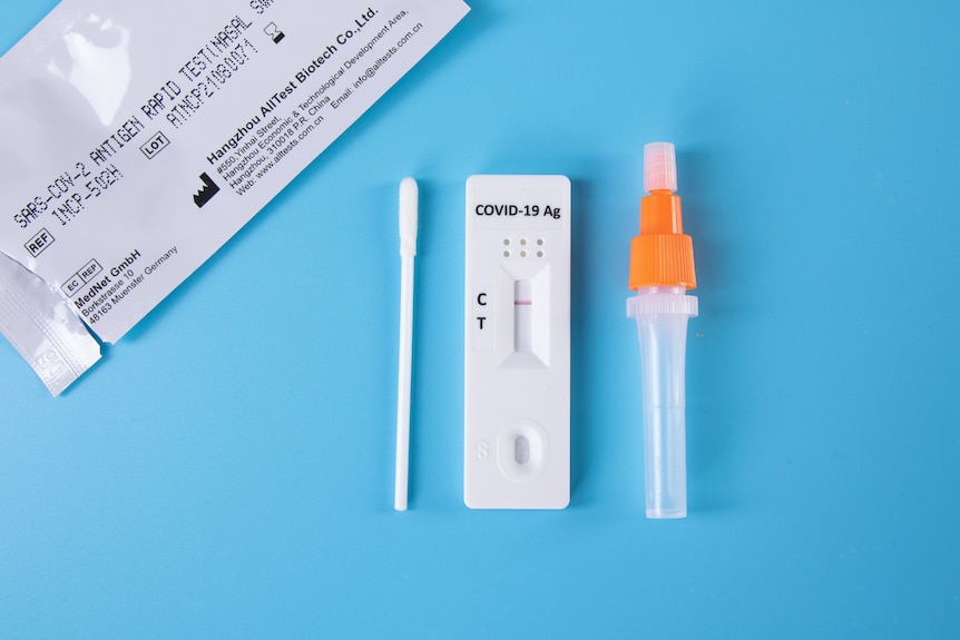 Descripción general de un paquete de prueba rápida de antígeno, un hisopo, un frasco gotero pequeño y la prueba misma colocada en una superficie azul.