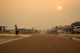 A man checks his phone on a driveway in a residential street, with the street covered in smoke haze from a bushfire.