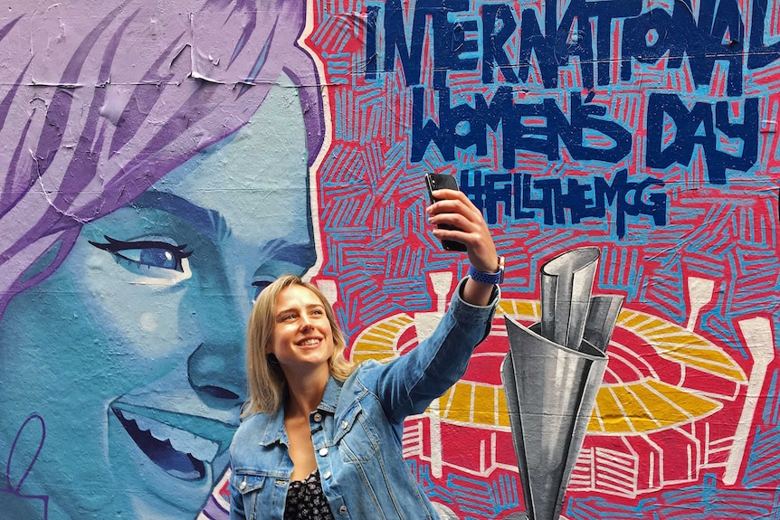 Ellyse Perry holds her phone above her head and looks to it to take a selfie.