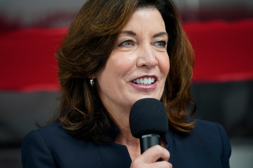 A woman in a suit jacket smiles as she raises a microphone to her mouth with her right hand.