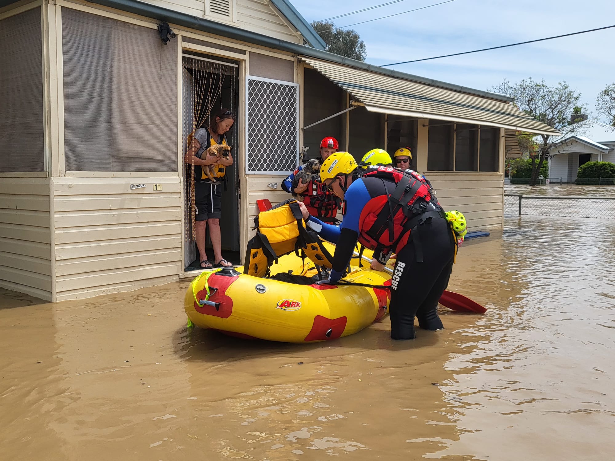 A resident standing in thongs in a door of a house with a dog about to get into a rescue raft in floodwaters with personnel.