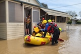 A resident standing in thongs in a door of a house with a dog about to get into a rescue raft in floodwaters with personnel.