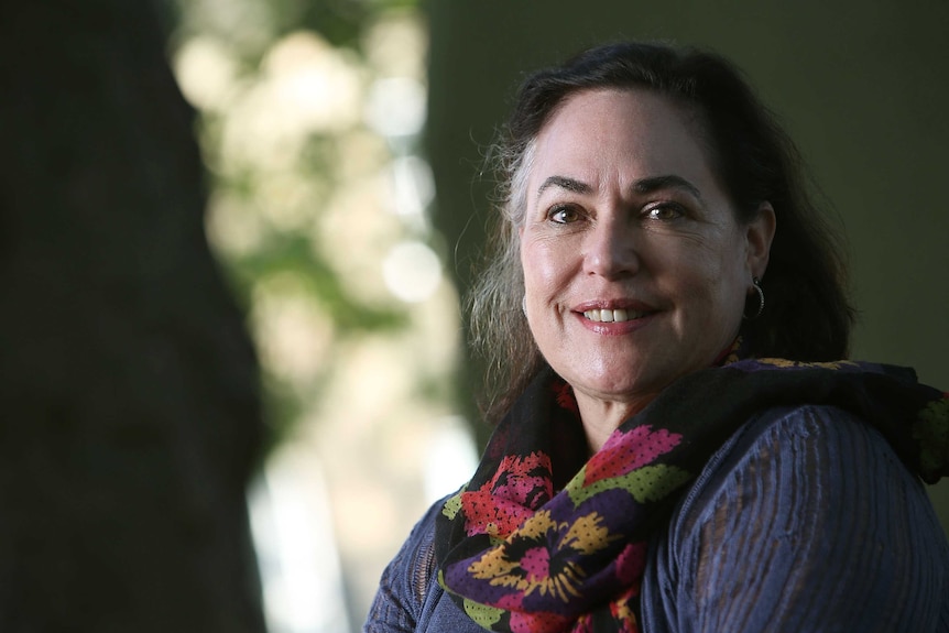 A portrait of bloom, a woman in her fifties, from the chest up: she smiles into the camera with a colourful scarf on her neck.