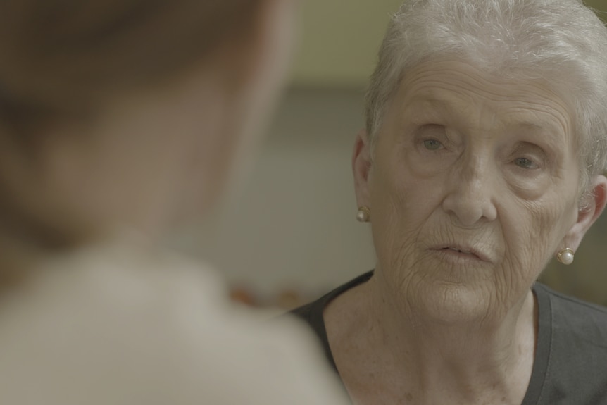 A woman with grey hair sitting at a table talking to another woman sitting opposite who is only partially visible in the photo.