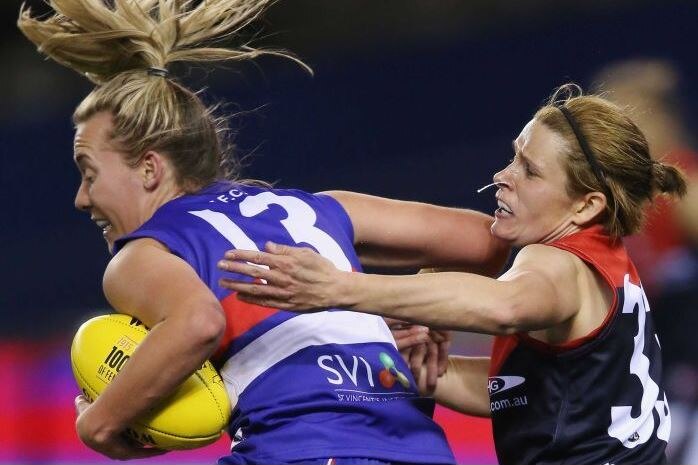 woman AFL with ball on left being tackled around shoulders from player on right from behind