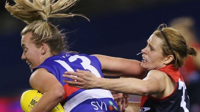 woman AFL with ball on left being tackled around shoulders from player on right from behind