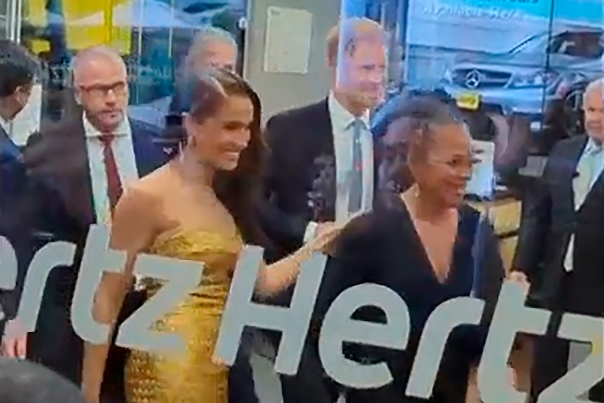 A still from a social media window shot through a window shows Meghan in gold dress alongside her mother and Prince Harry.