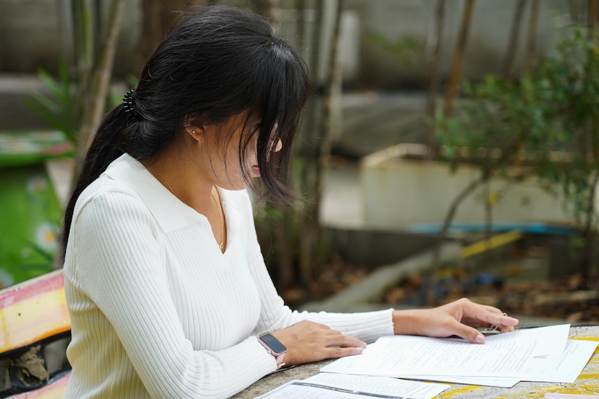 A woman wearing a white long-sleeved top, whose hair covers her face, sits at a table looking at documents