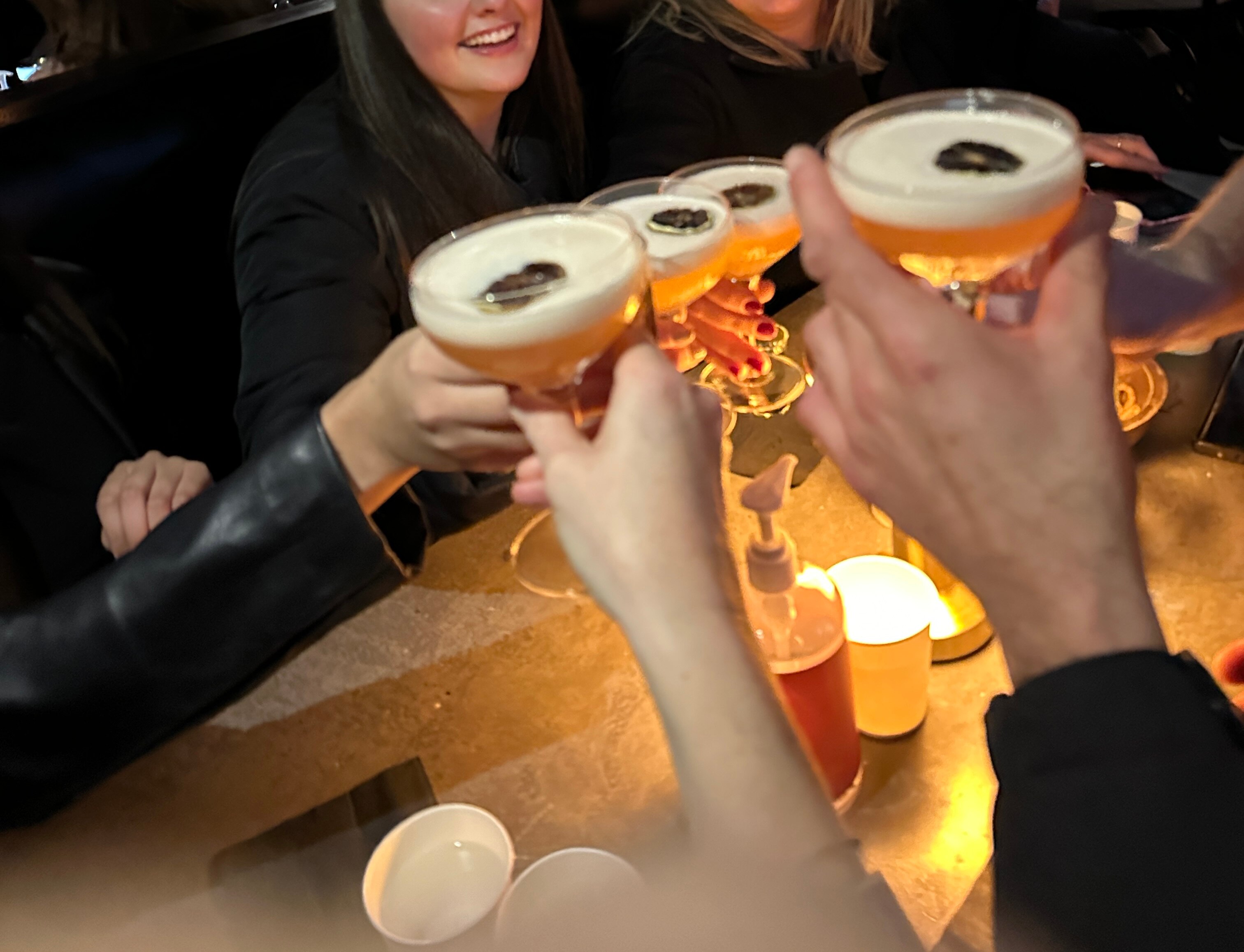 Four cocktail glasses are raised to touch in a cheers gesture over a table 