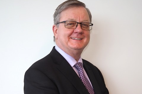 James O'Halloran looks to the camera in a mid-shot. He wears a black suit, a light shirt and purple and pink tie and has glasses