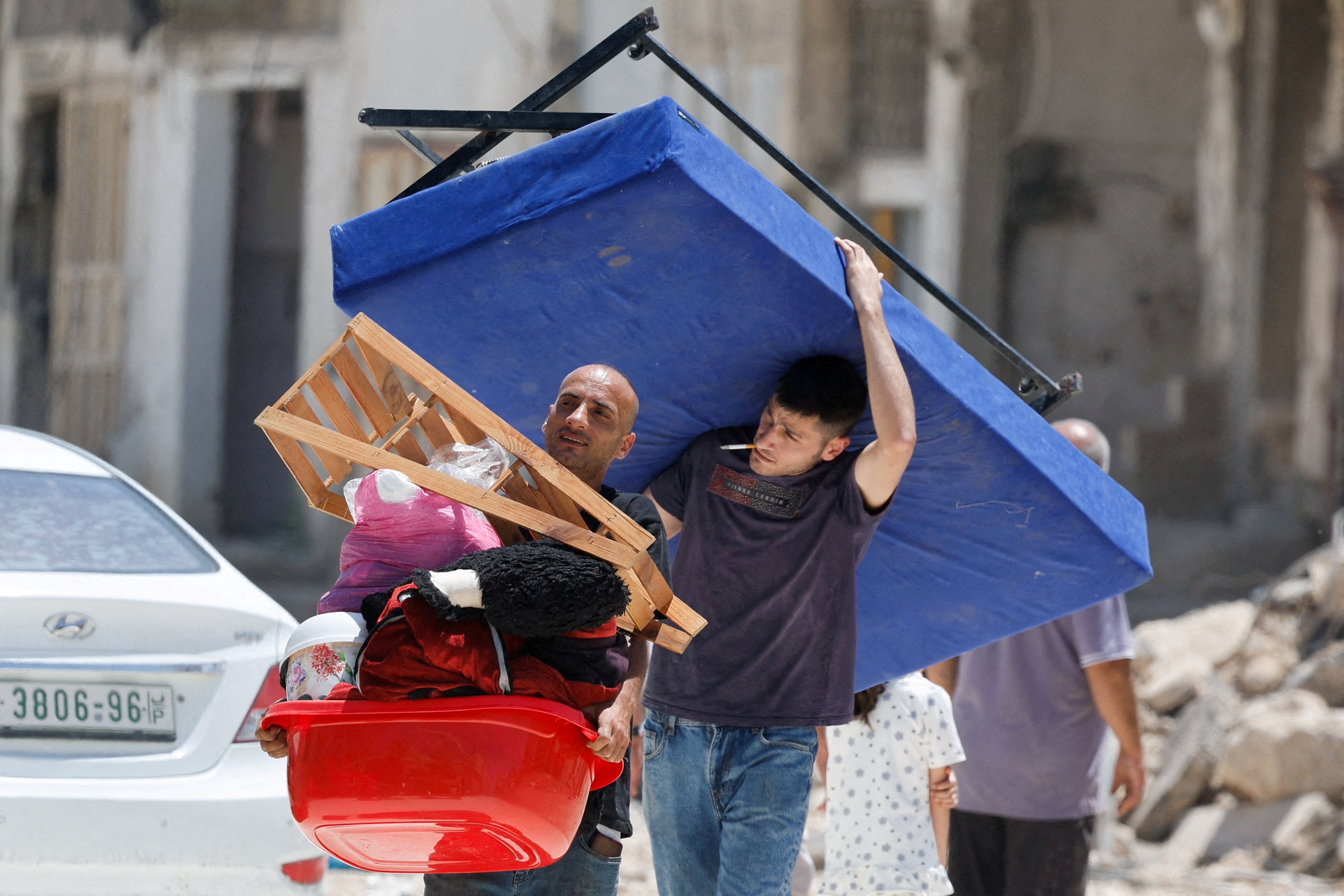 Two Palestinian men carry a table and their belongings as they walk near rubble.
