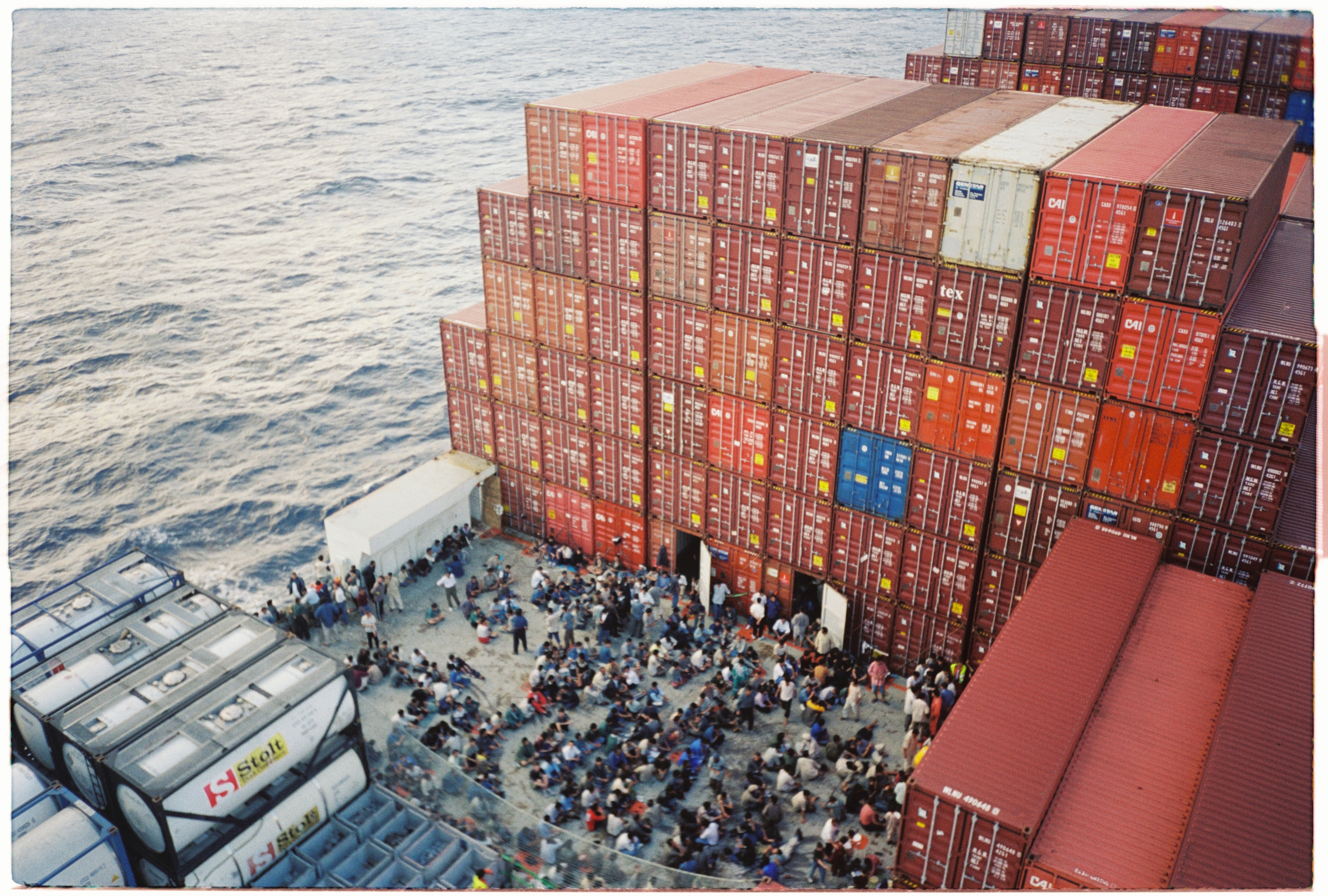 An overhead shot of a crowd of people sitting and standing on the deck of a large freight ship.