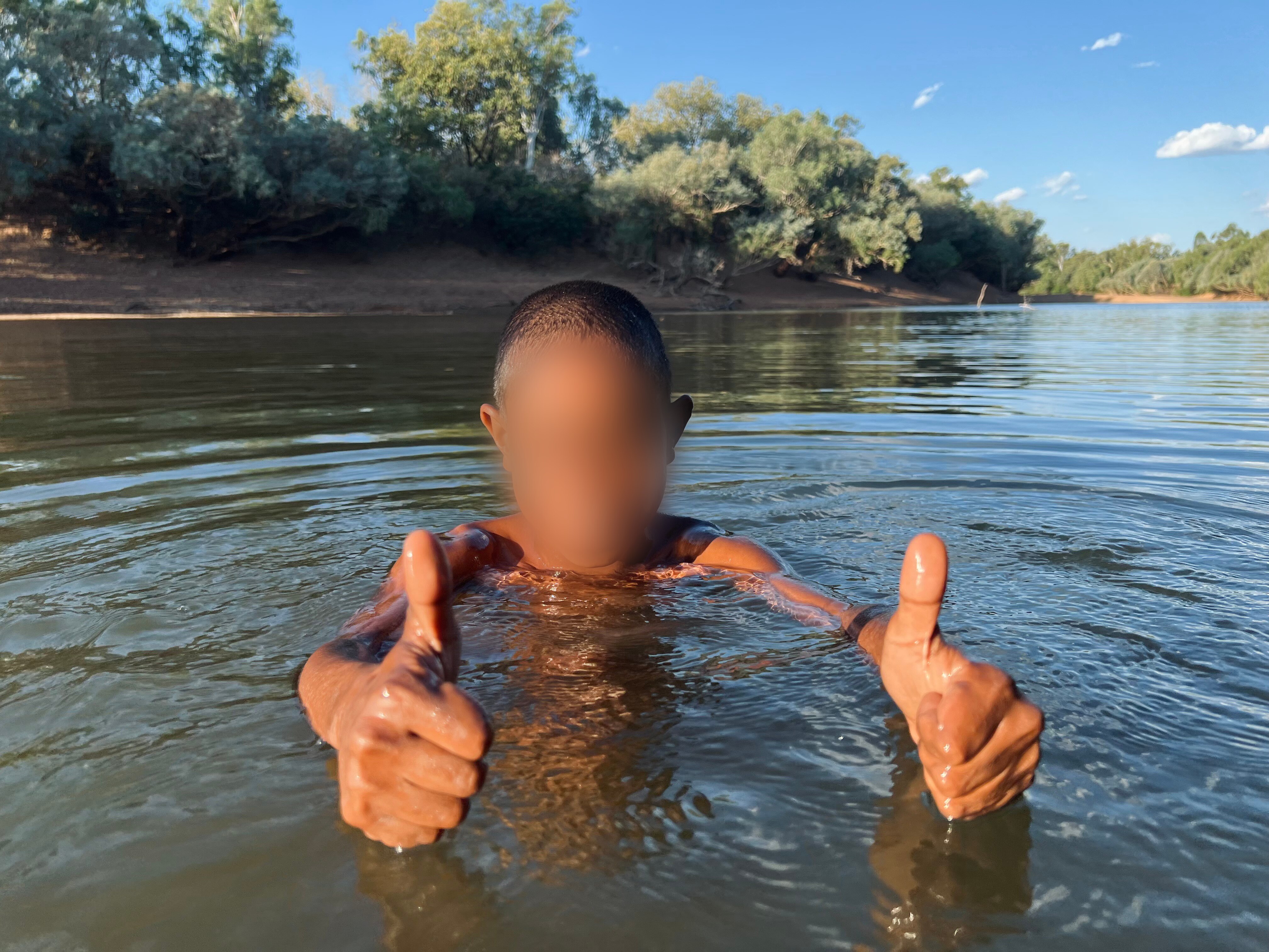 a young boy with a pixelated face gives thumbs up while swimming in a river
