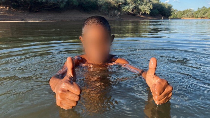 a young boy with a pixelated face gives thumbs up while swimming in a river