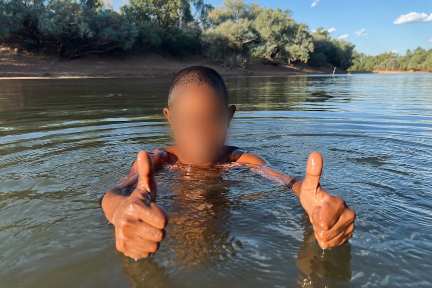 a young boy with a pixelated face gives thumbs up while swimming in a river