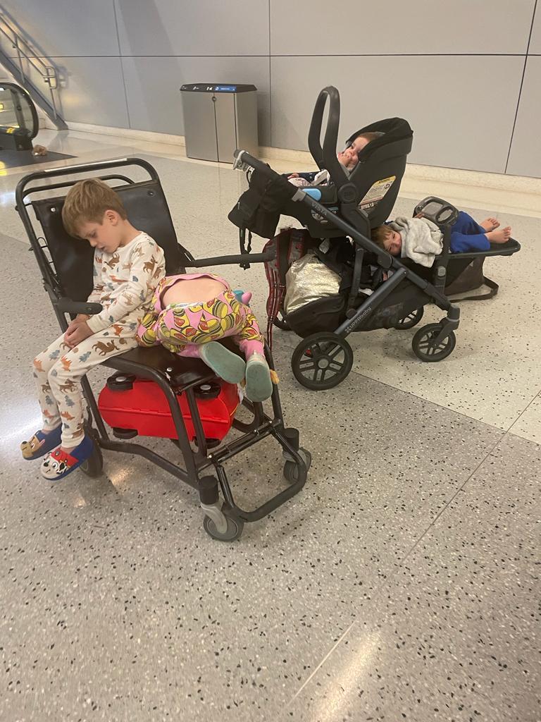 Four children sitting on two chairs, including a pram, inside an airport terminal.