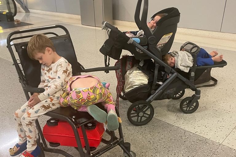 Four children sitting on two chairs, including a pram, inside an airport terminal.