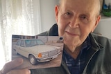 Old man looking at camera, holding up photograph of cream Mercedes Benz car.