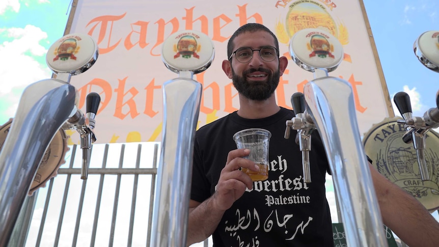 A bespectacled bearded man standing behind a beer tap holds a cup containing amber liquid
