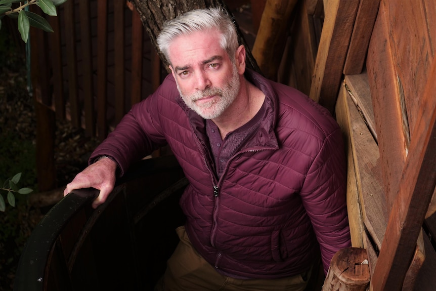 Portrait of a man with grey hair and grey beard, wearing a maroon puffer, in treehouse staircase.