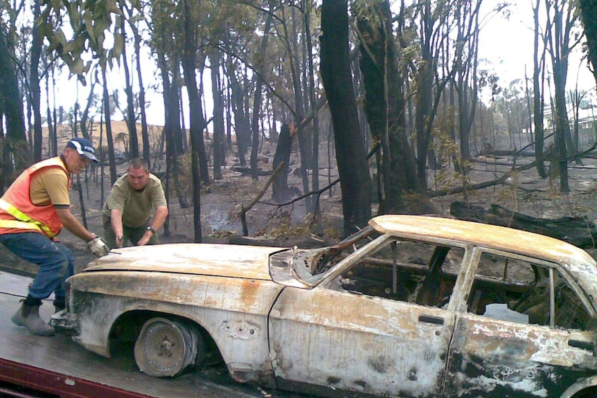 Two men pull a burnt out shell of a car on a tow truck