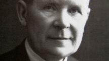 A black and white photo of a man in a black suit and striped black tie.