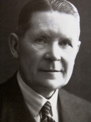 A black and white photo of a man in a black suit and striped black tie.