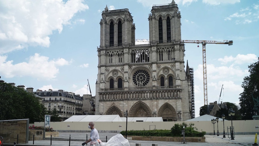 A man in white working gear drags a large white bag in front Notre Dame, with scaffolding and cranes visible.