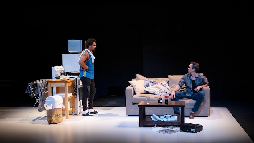 Two dark-skinned men in conversation on a stage set, one standing by an oven, the other sitting on a couch.