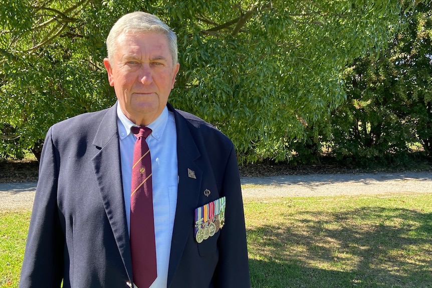 An elderly man stands with in front of a tree with a blue blazer on, with four gold medals attached to the front of his blazer.