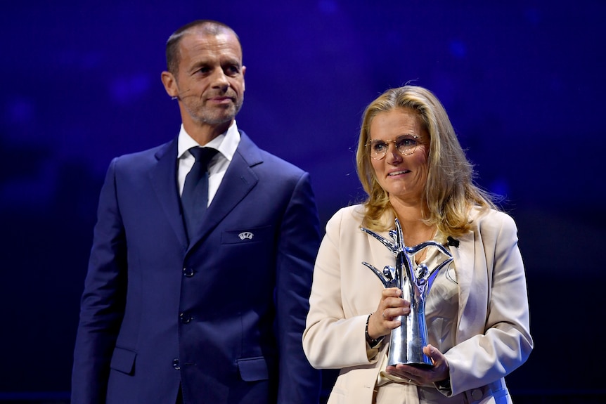 A football executive wearing a headset microphone watches on as England's women's football coach holds a trophy.