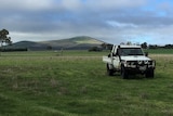 a ute drives through a grassy paddock, in the background there are rolling hills, some covered in clouds