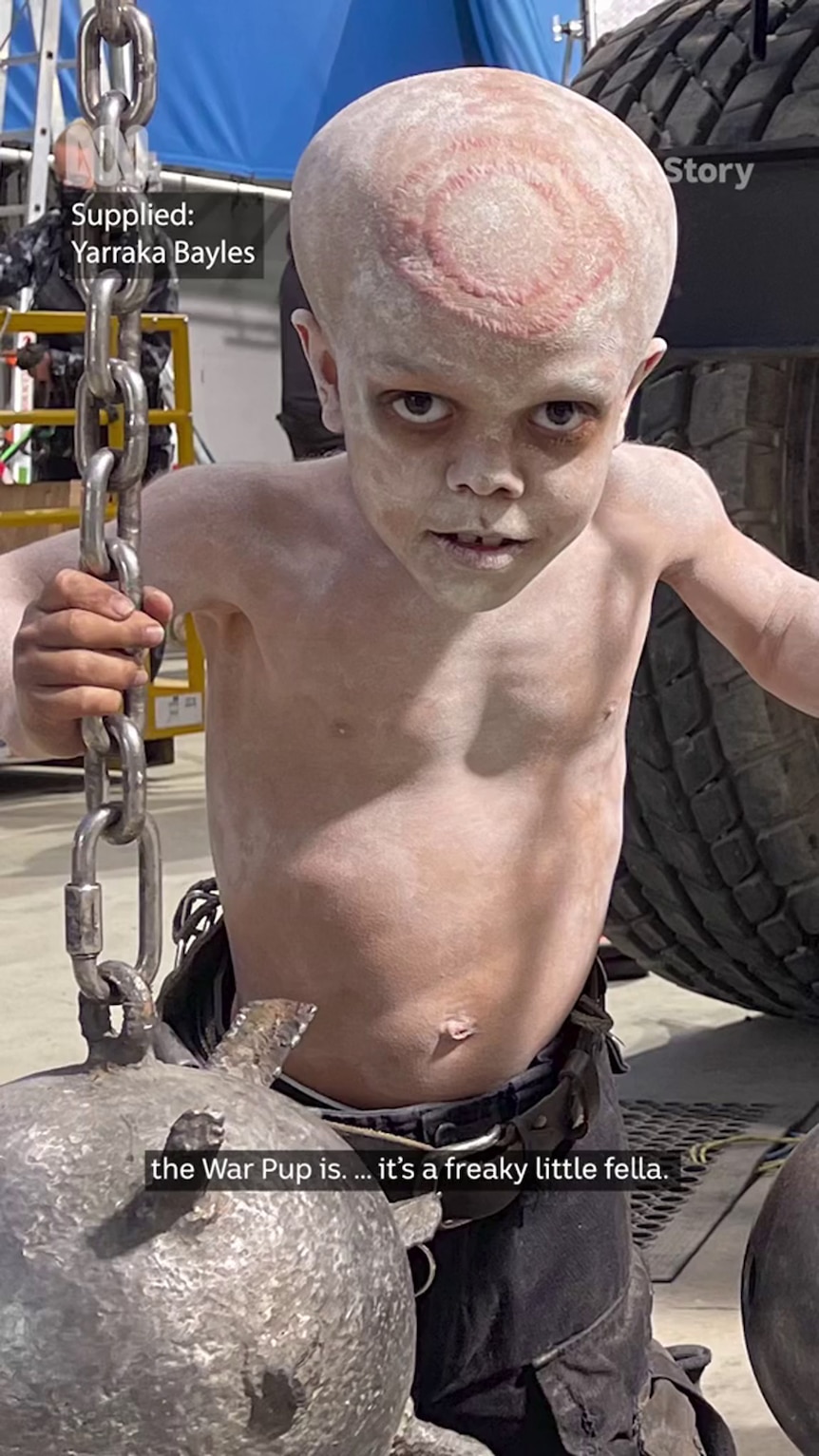 A shirtless boy with medium-tone skin has body paint, including a circular design on his head, and he holds onto a chain