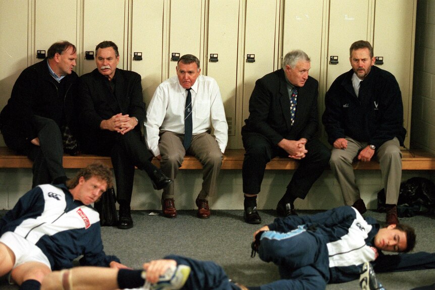 A group of Victorian coaches sit on a bench in the change rooms before a State of Origin, while players stretch.  