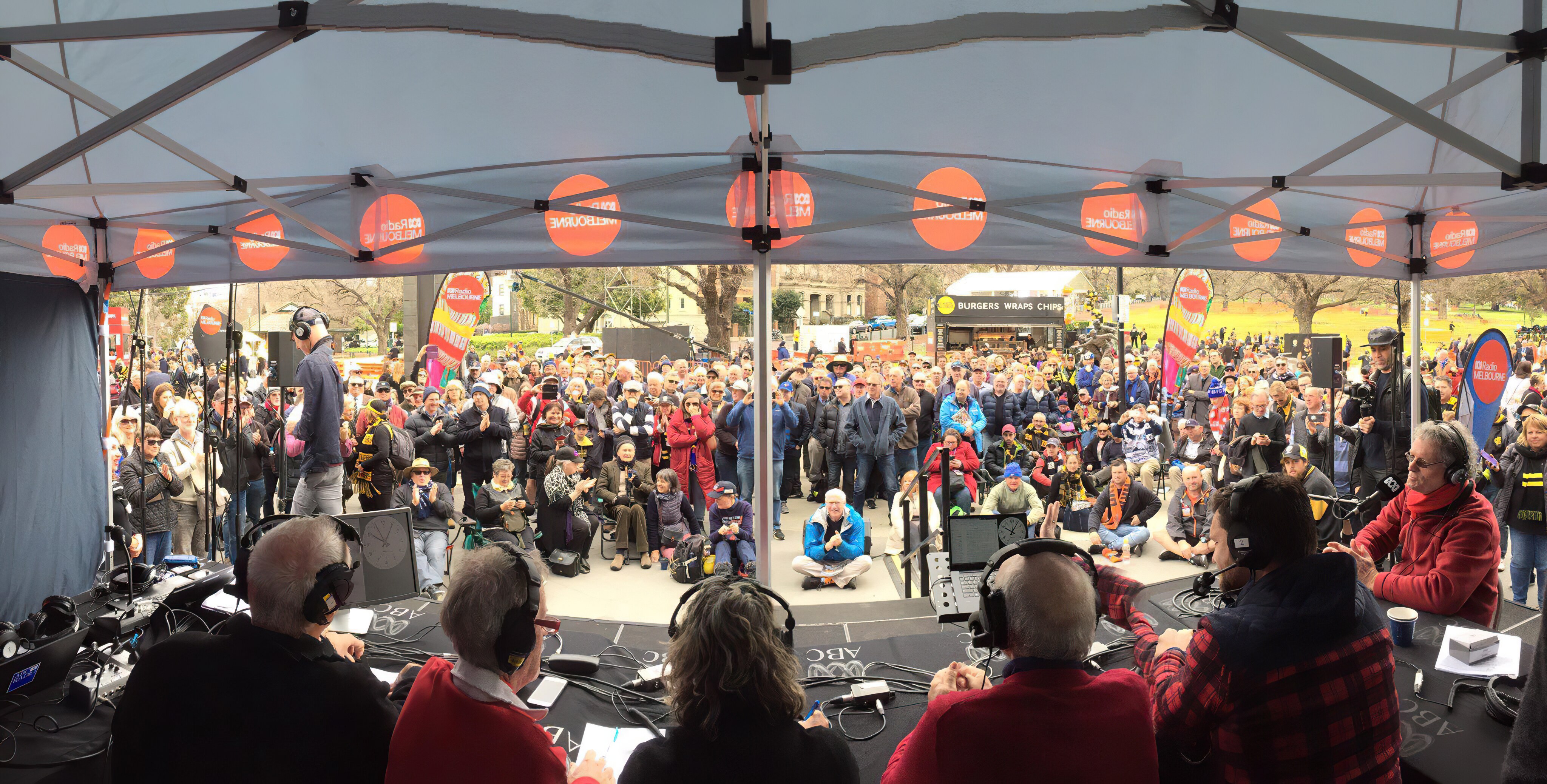 Backs of six men sitting at radio desk in front of microphones looking out at large crowd watching.