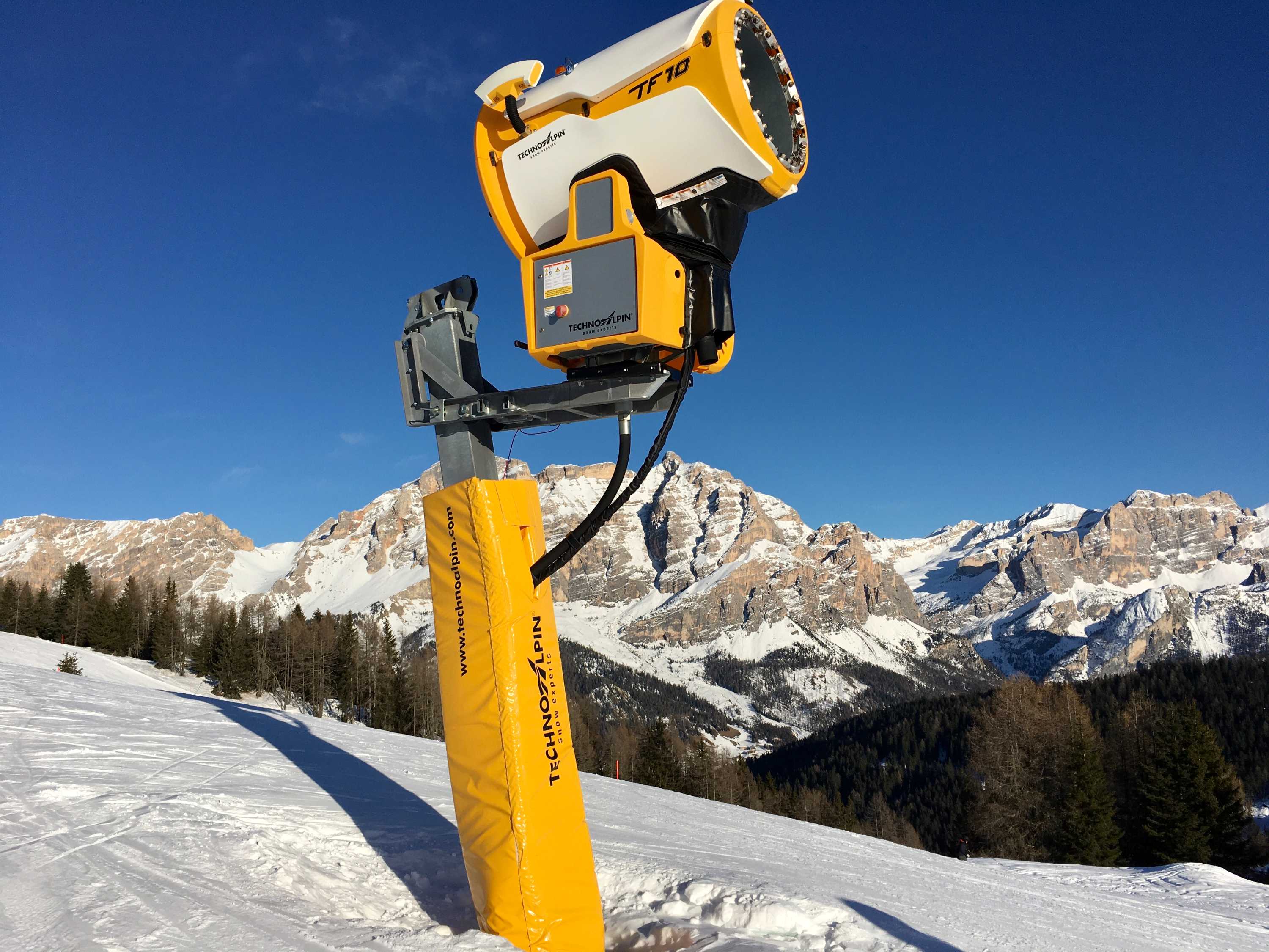 A yellow snow cannon with snowy mountains in the background.