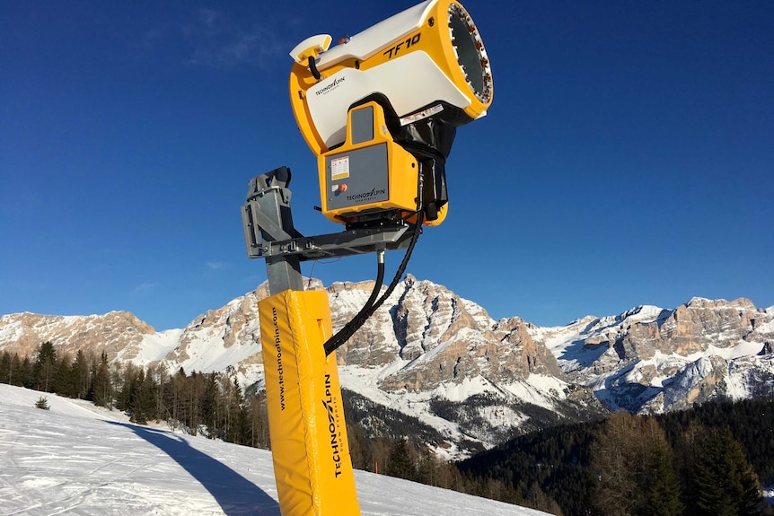 A yellow snow cannon with snowy mountains in the background.