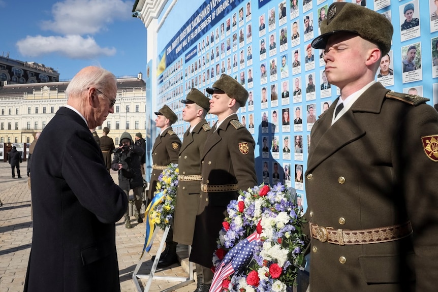 Biden holds his hand over his heart as he looks towards a wreath leaning against a large wall displaying photos of soldiers