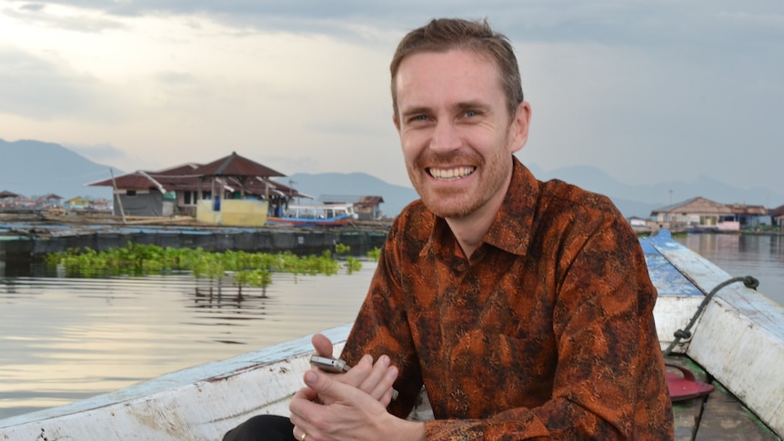 A man wearing an orange and brown paisley shirt smiles at the camera while sitting in a boat on a river