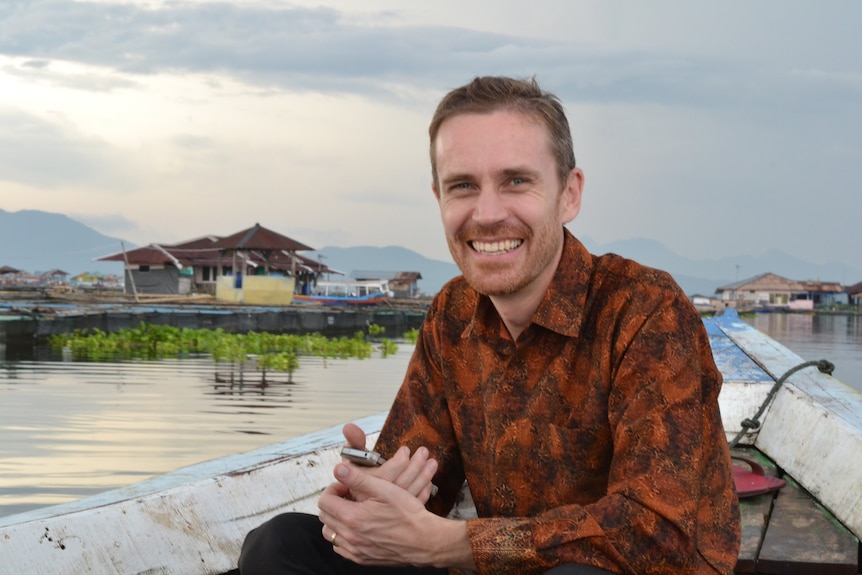 A man wearing an orange and brown paisley shirt smiles at the camera while sitting in a boat on a river