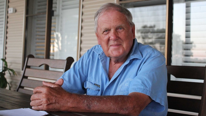 An older man sits with his hands clasped at a table on a veranda, smiling.