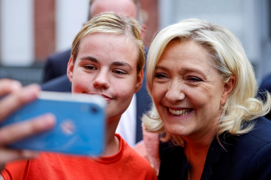 Marine Le Pen smiles as a young woman next to her extends her phone in front of them to take a selfie