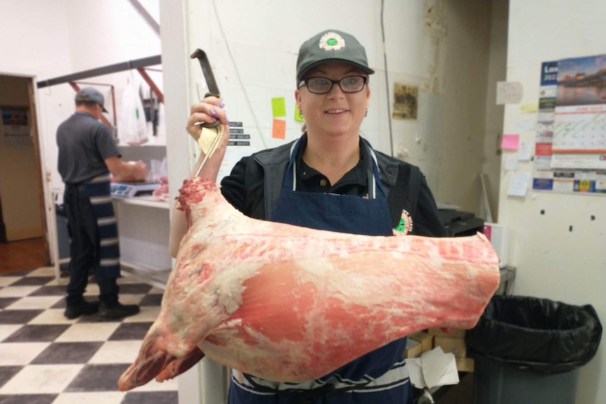 A woman smiles, wearing dark clothes and an apron. She holds a skinned animal carcass and stands in an industrial butchers room