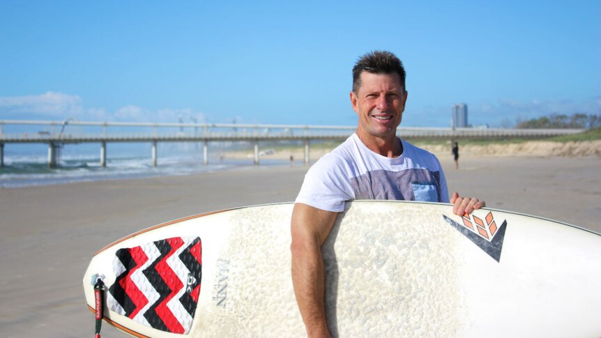 A man wearing a white t-shirt holding a surfboard, standing on a Gold Coast beach.