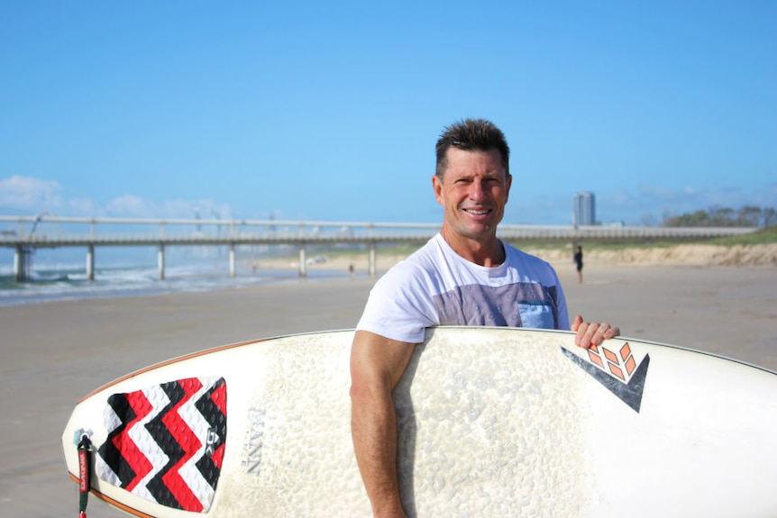 A man wearing a white t-shirt holding a surfboard, standing on a Gold Coast beach.