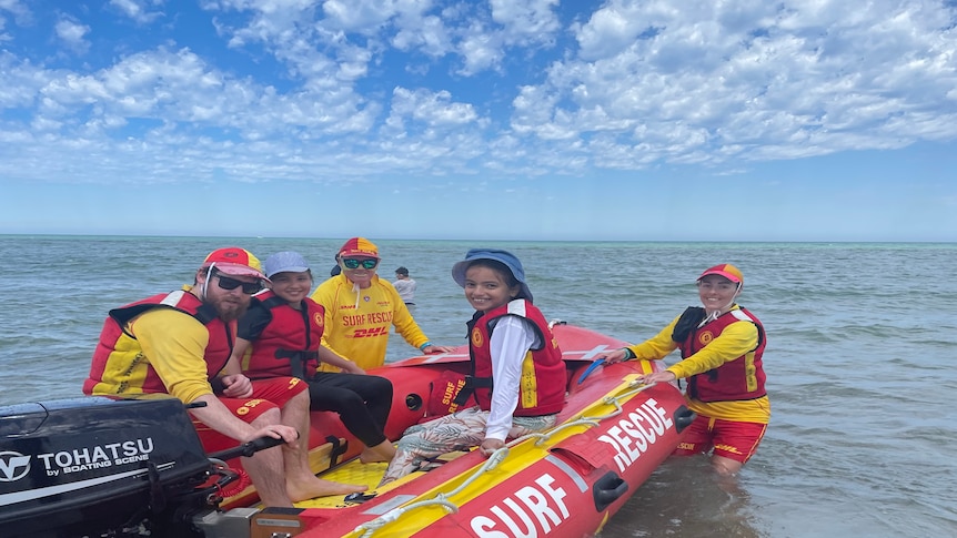 Two children sit in a surf life saving boat in between three surf life savers. The boat is in the ocean 