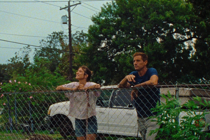 A brown-haired woman smiles widely as she leans on a wire fence beside a sombre-looking 40-something man smoking a cigarette