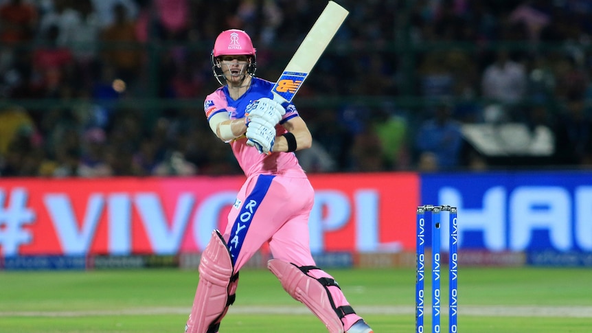 Steve Smith holds his bat up and looks behind him wearing pink cricket clothes.